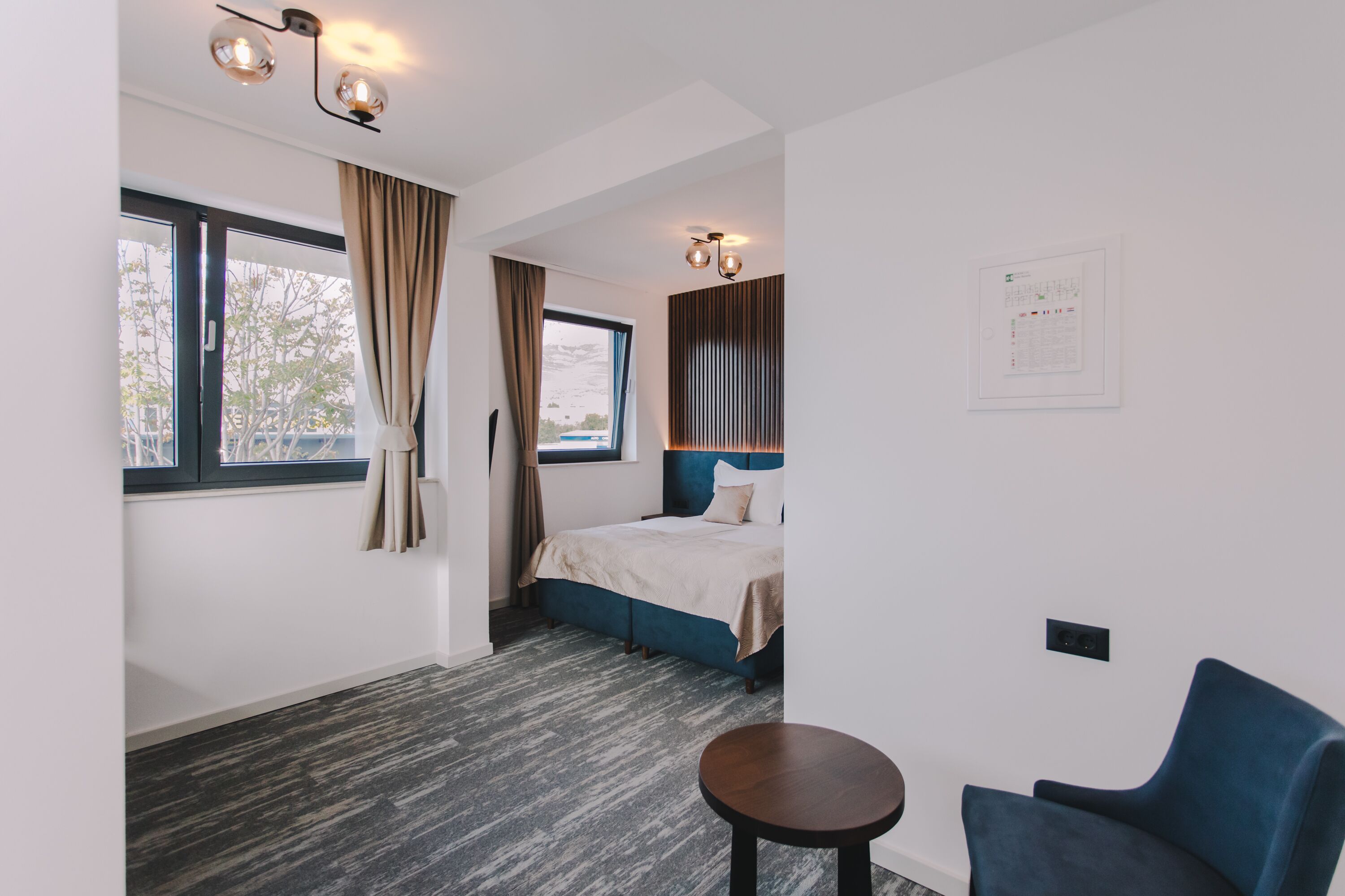 Quarto de hotel moderno com uma cama grande, uma mesa redonda pequena e uma poltrona azul.