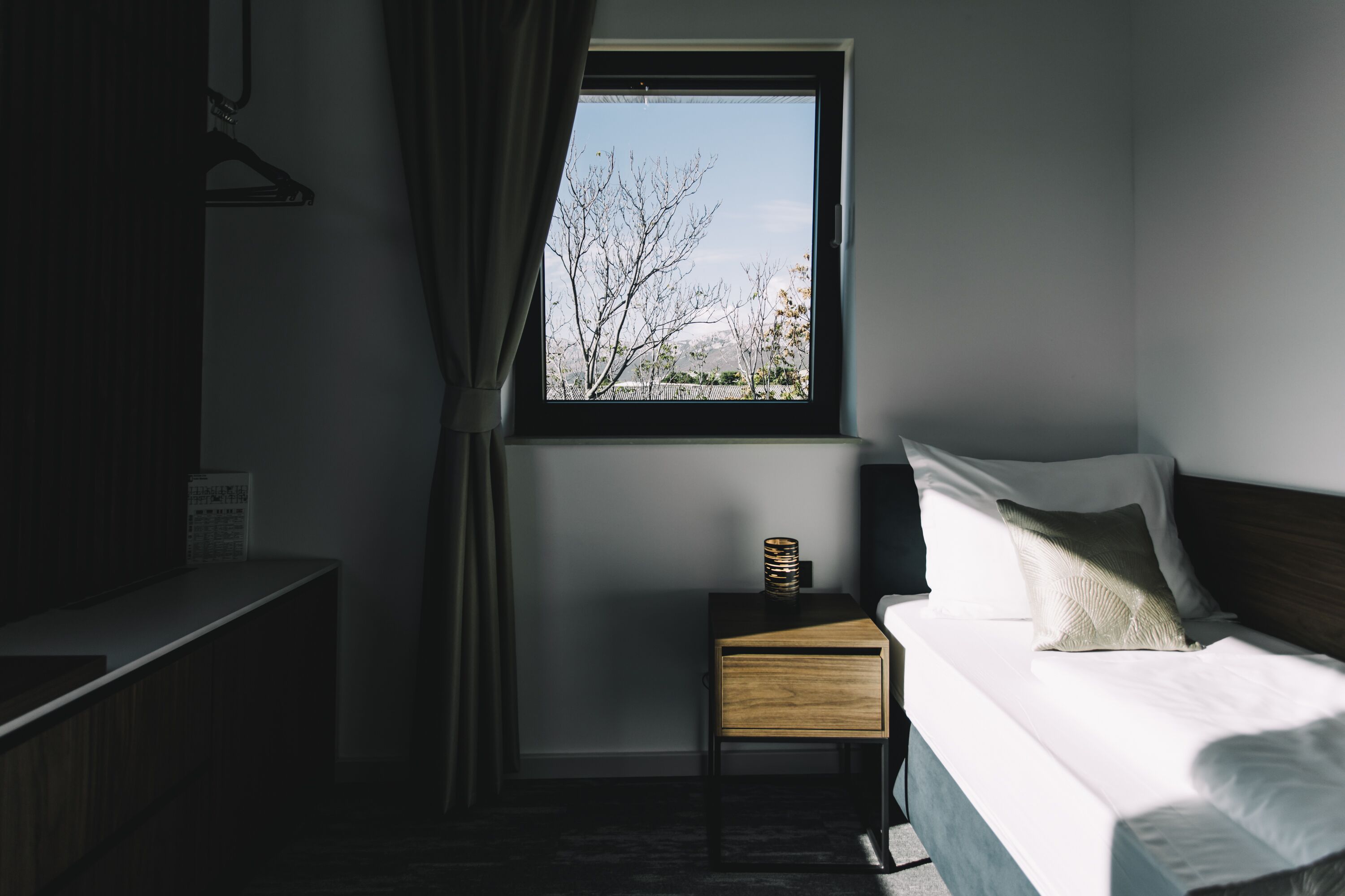 Habitación de hotel con cama, mesita de noche, ventana con vistas a árboles y cortina.