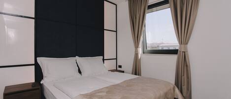 A modern hotel room with a large bed, a bedside table, a nightstand, and a window with curtains.