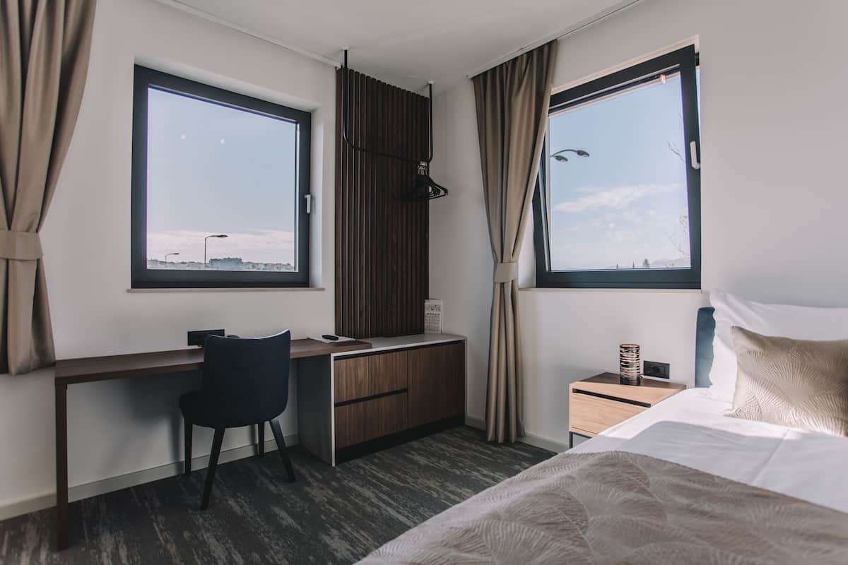 A hotel room with a bed, a desk, and two windows.