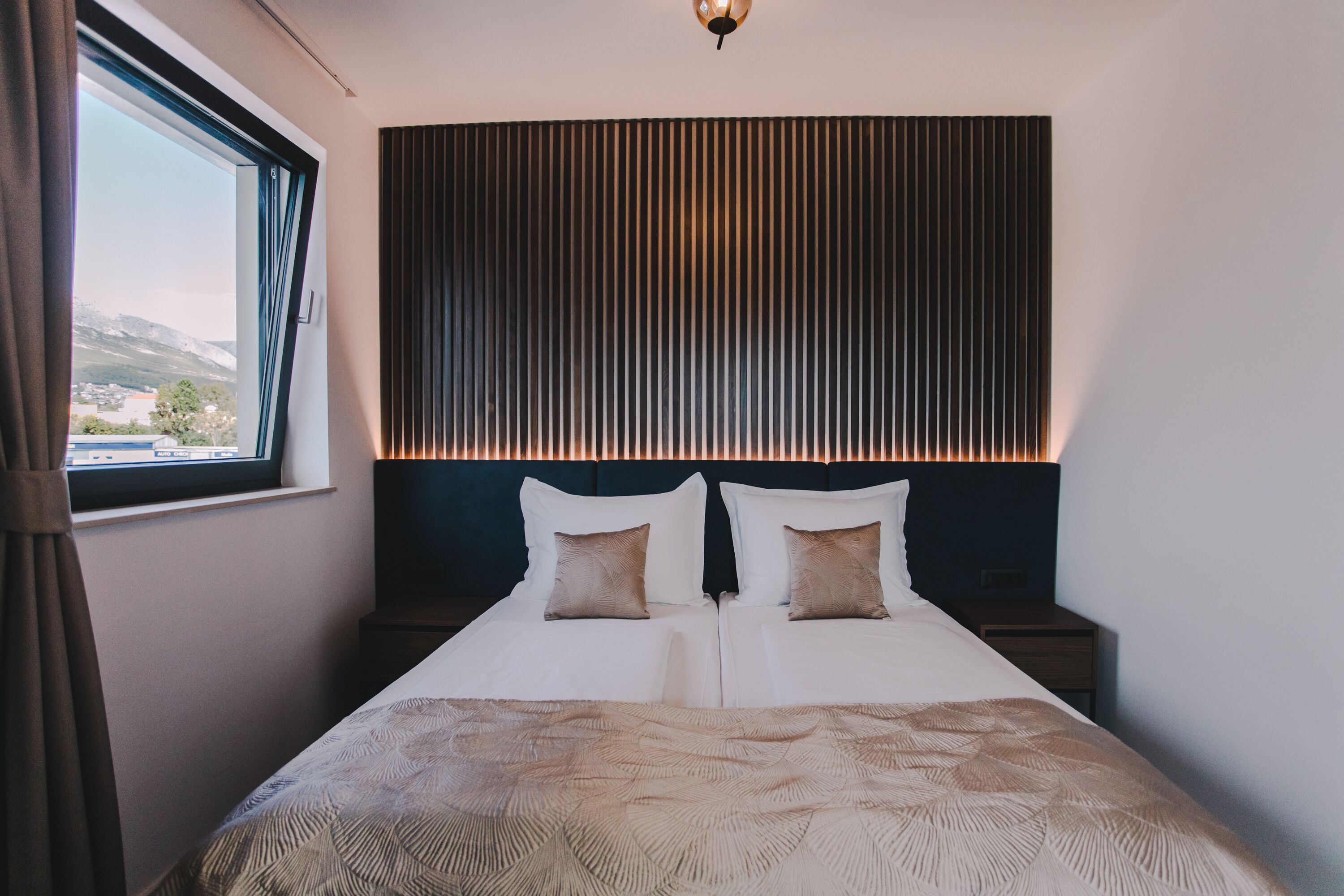 Una habitación de hotel moderna con una cama grande, un cabecero con listones verticales y vistas a la montaña a través de una ventana.
