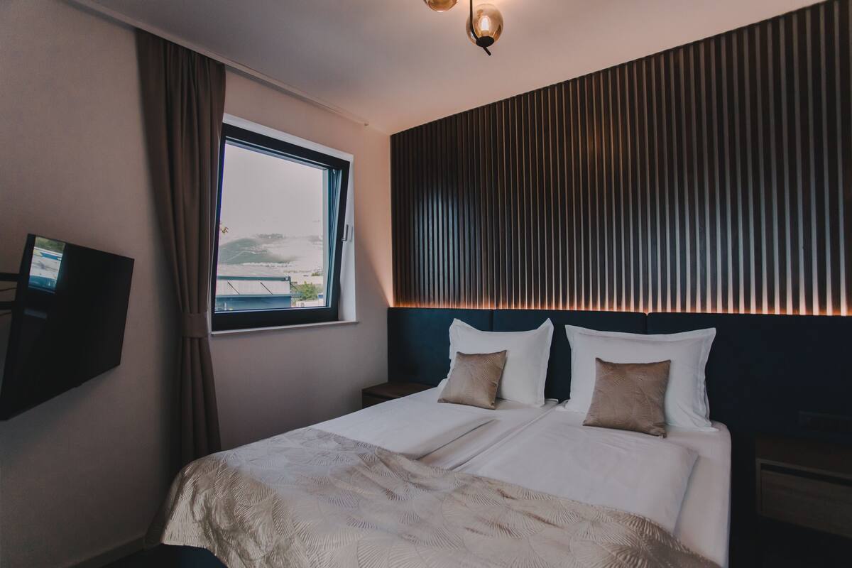 A hotel room with a bed, a TV mounted on the wall, a window with curtains, and a view of the sea.