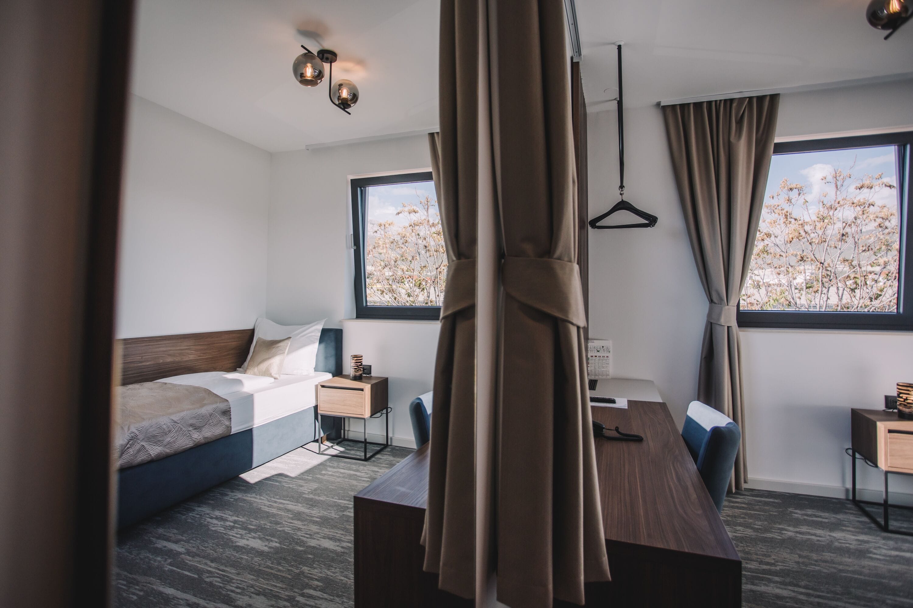 Una habitación de hotel moderna con cama, escritorio y silla. Hay dos ventanas con cortinas, una lámpara colgante y una pequeña mesita de noche.