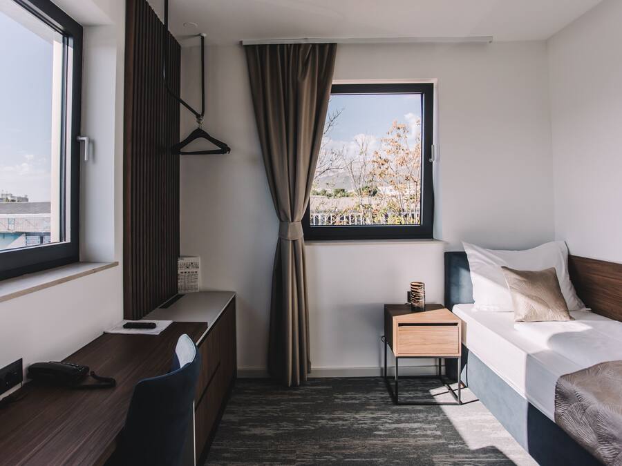 A hotel room with a bed, a desk, a chair, a small table, a window with a view of buildings, and a curtain.