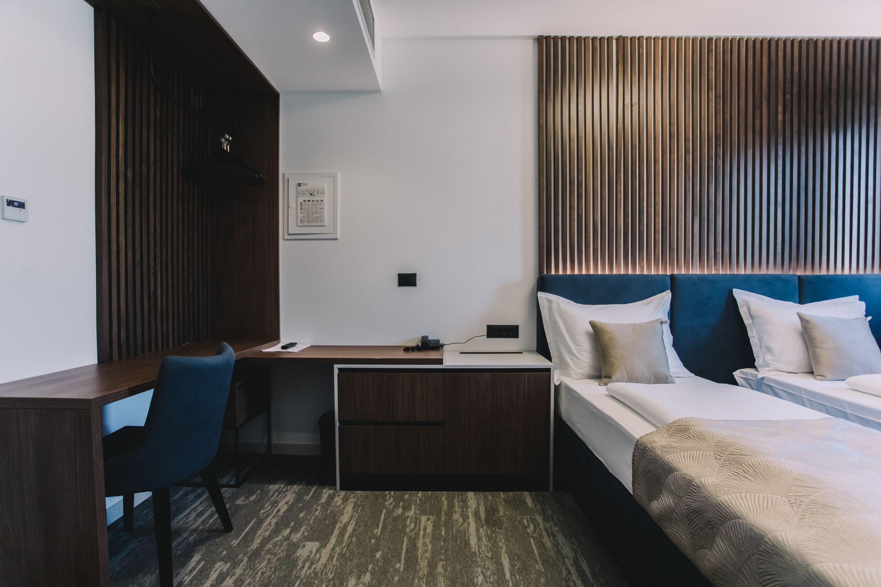 Quarto de hotel com escrivaninha, cama com cabeceira azul e design de parede com ripas de madeira.