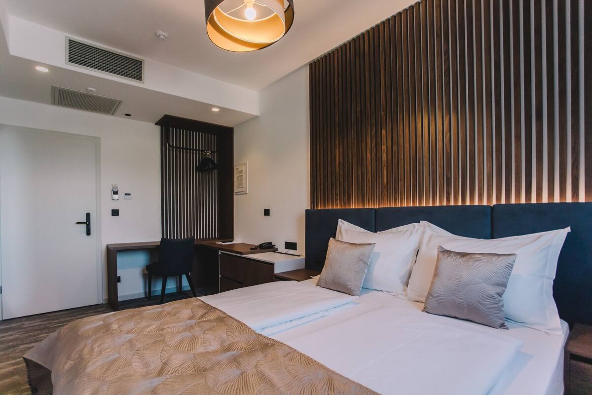 A hotel room with a large bed, a desk, and a chair. There is a wooden panel wall and a ceiling light fixture.