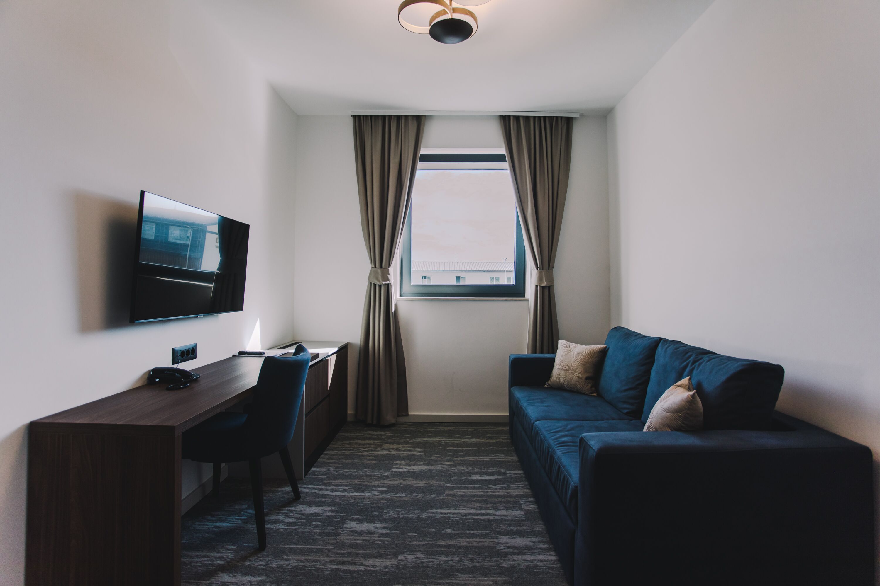 Ein Hotelzimmer mit dunklem Holzschreibtisch, einem blauen Sofa, einem Fernseher und einem Fenster mit Vorhängen.