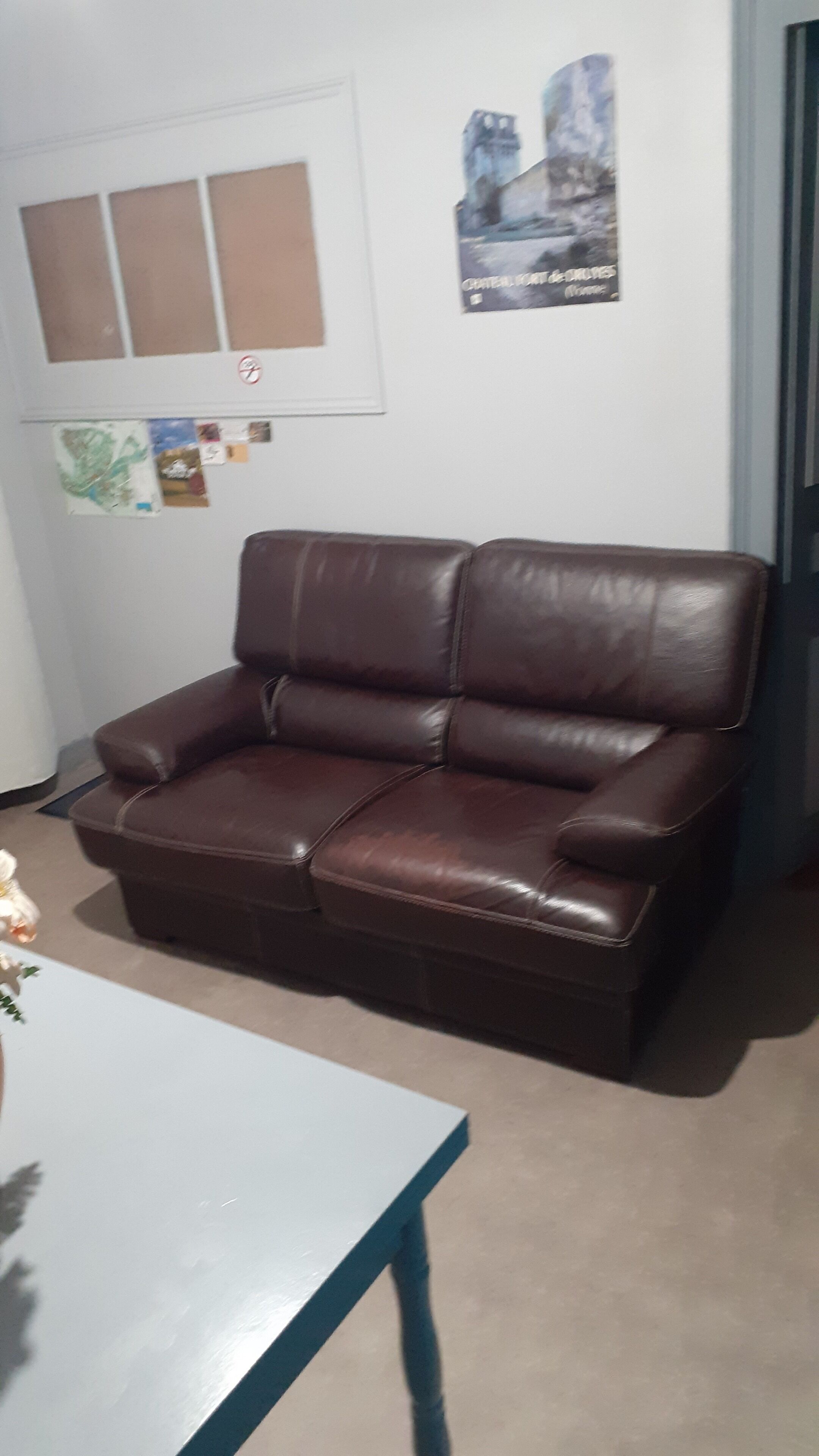 A brown leather sofa in a room with a white table, a window with blinds, and a poster on the wall.