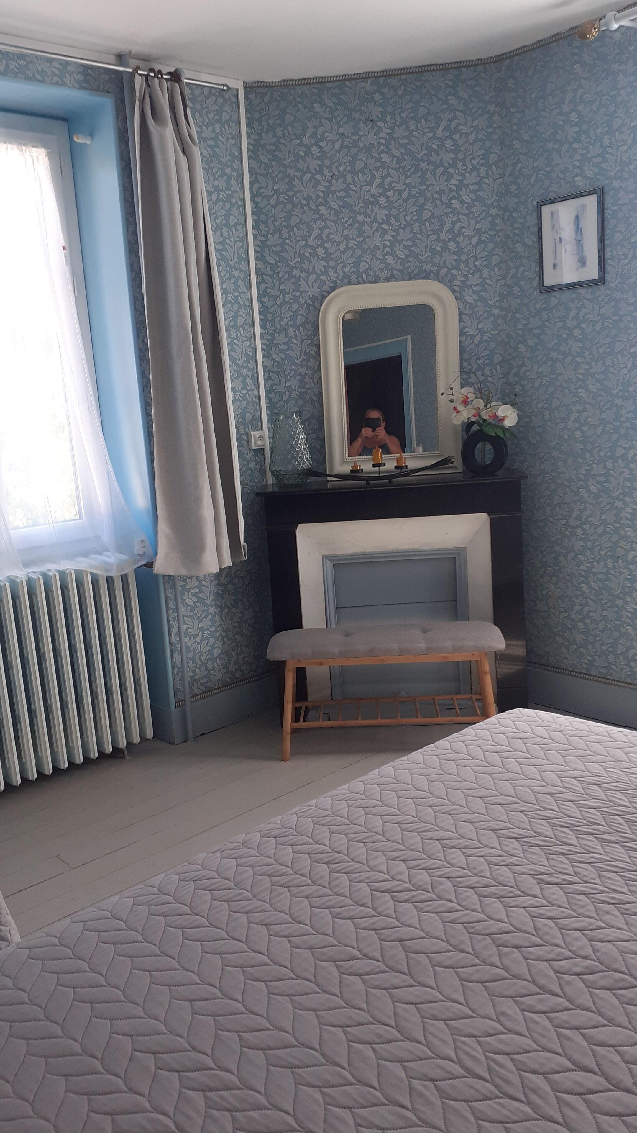 Ein Schlafzimmer mit einem Bett, einer Bank, einem Spiegel, einem Heizkörper und einem Fenster mit Vorhängen.