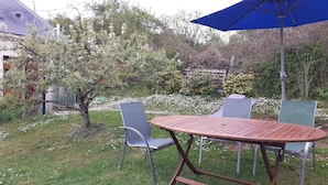 Un espace extérieur aménagé avec une table et des chaises, un parasol bleu et un jardin avec des arbres en fleurs.