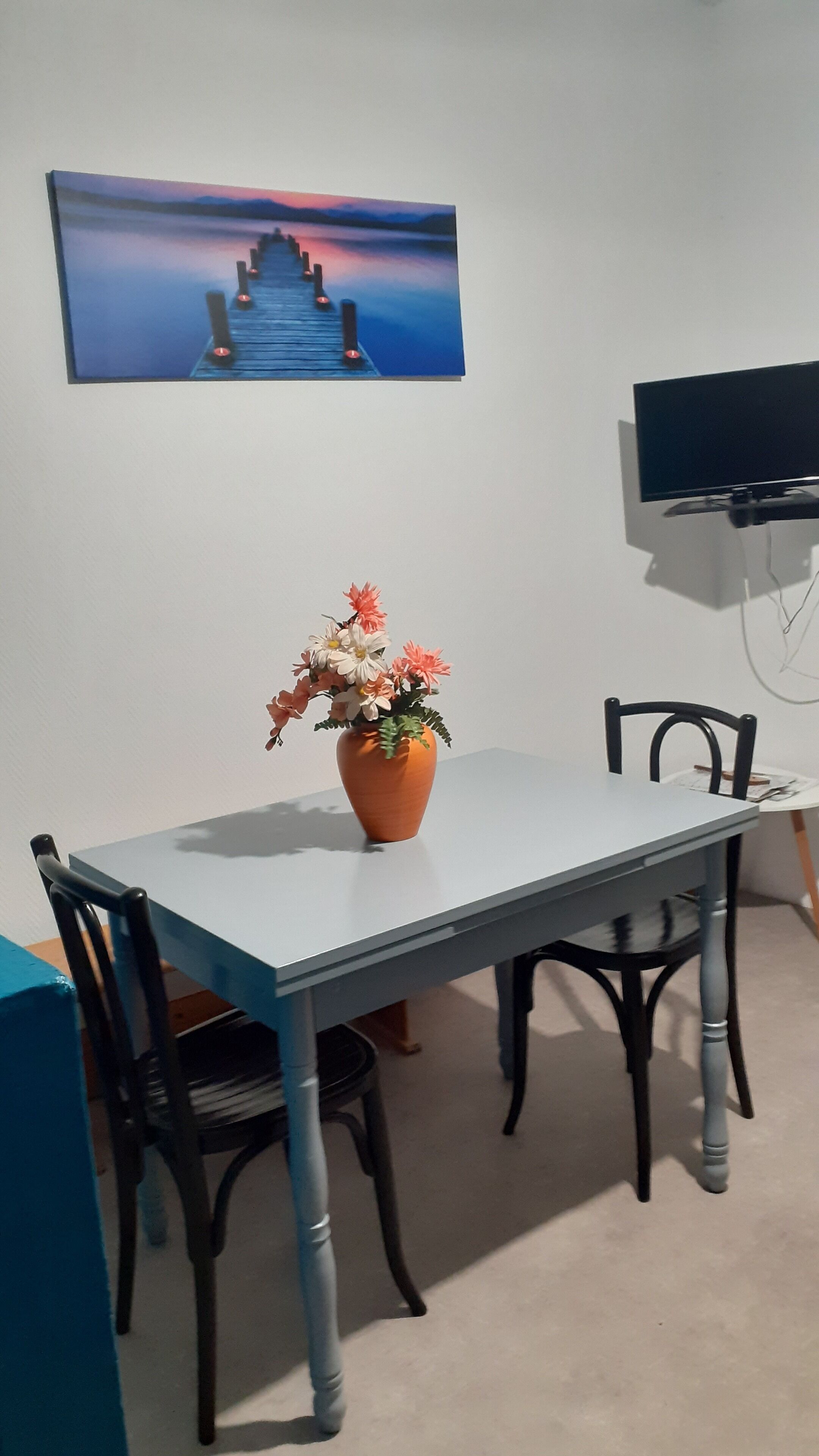 Un comedor con una mesa, sillas, un jarrón de flores, un cuadro y un televisor en la pared.