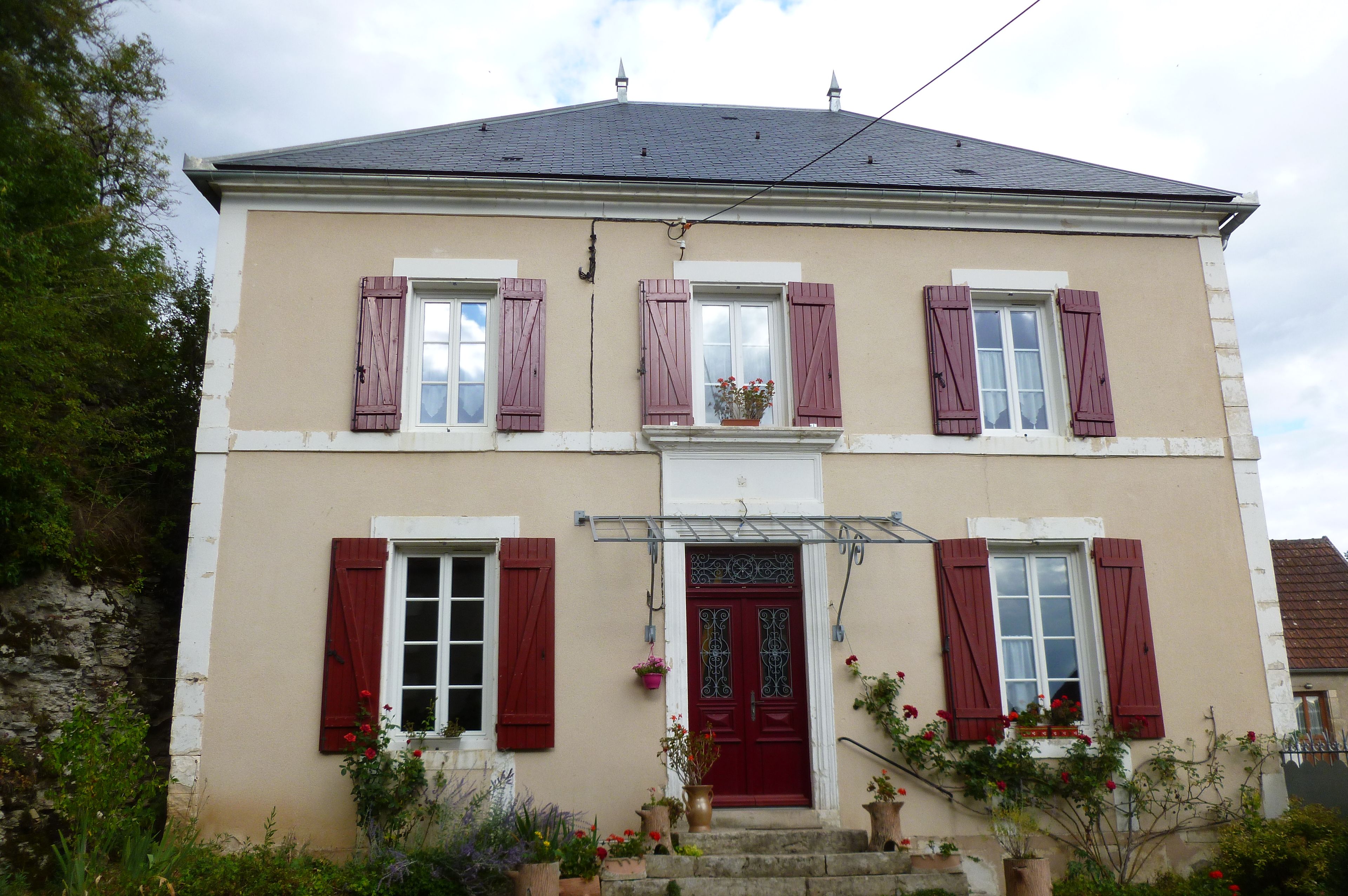 Un bâtiment à deux étages avec une porte rouge, de nombreuses fenêtres équipées de volets et un petit porche agrémenté de plantes en pot.