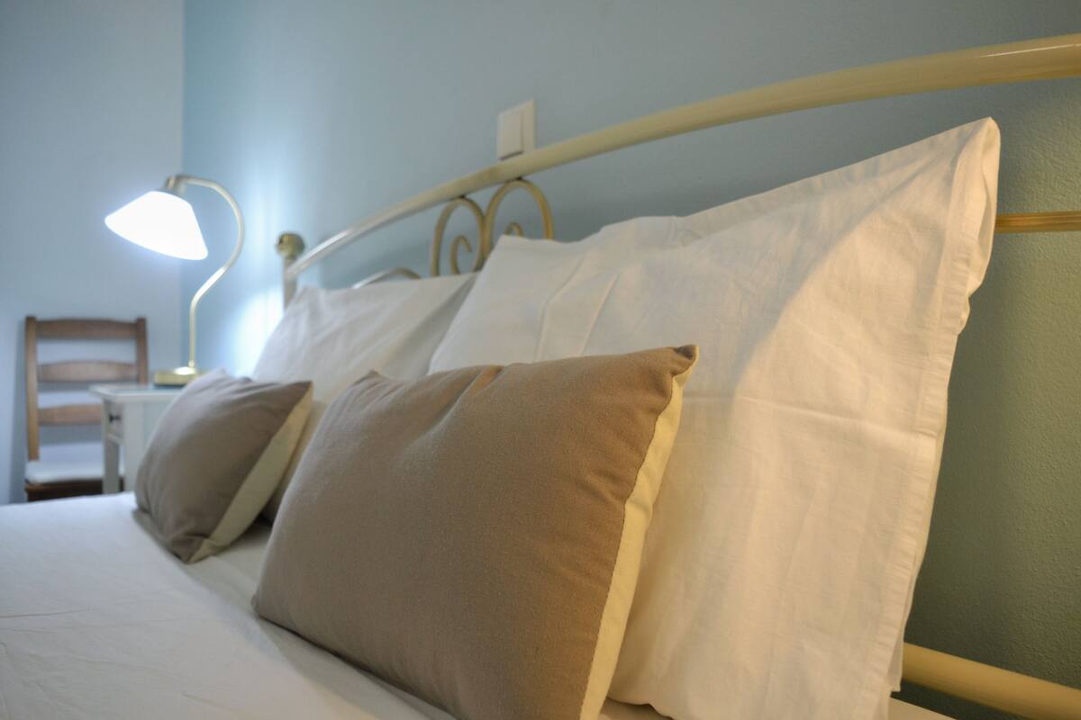 Uma cama com almofadas brancas e bege, uma cabeceira de metal e um abajur.