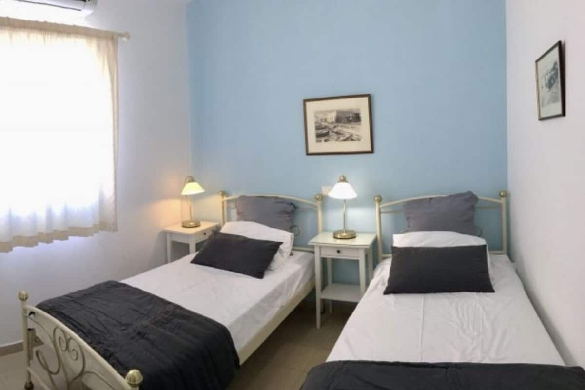 Quarto de hotel com duas camas de solteiro, televisão, janela com cortinas, mesa de cabeceira com abajur e um quadro na parede.