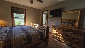 Une chambre avec un grand lit, une commode en bois, un téléviseur fixé au mur et une fenêtre donnant sur un paysage verdoyant.