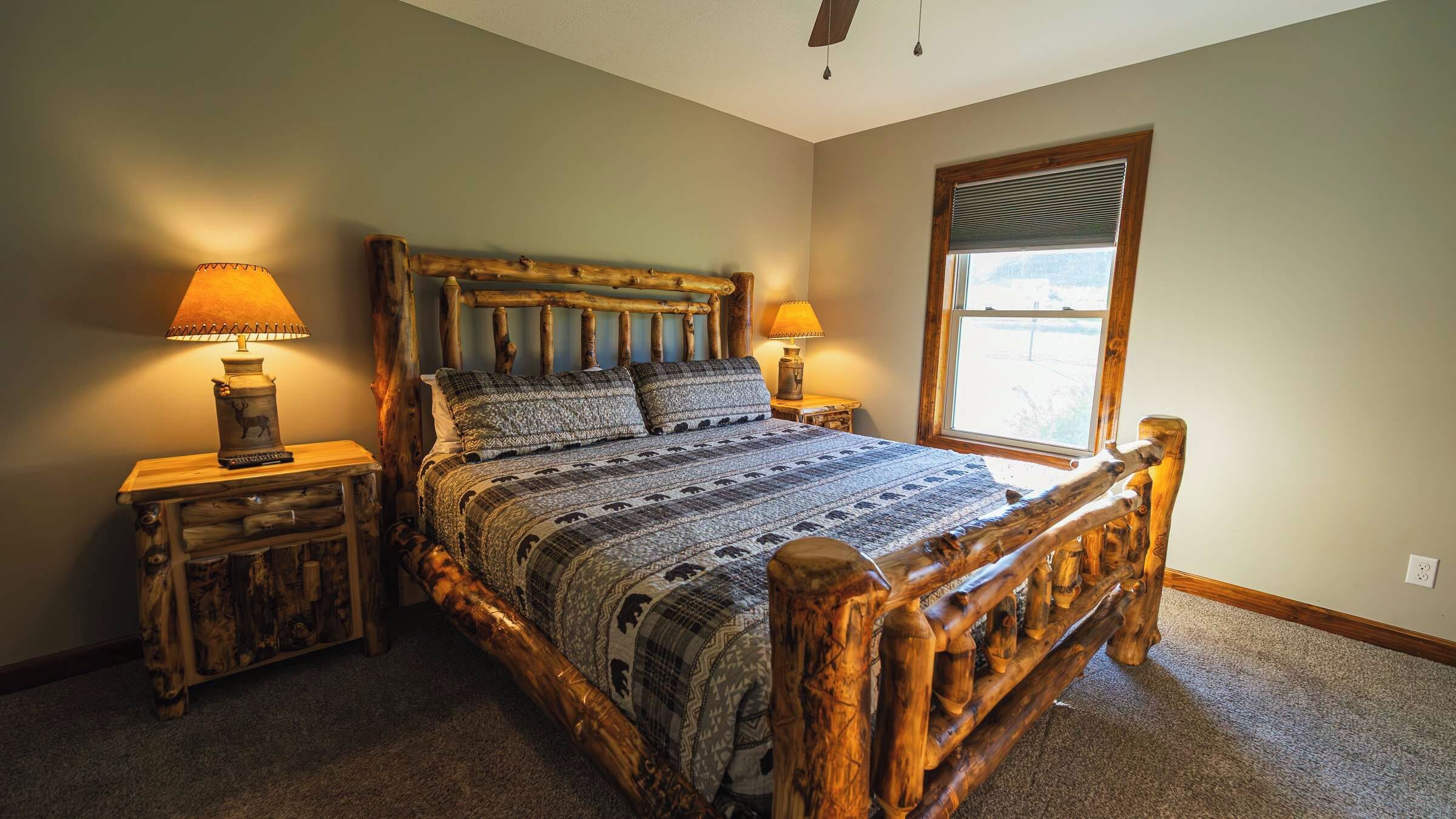 A bedroom with a wooden bed frame, two bedside tables with lamps, a window with blinds, and a carpeted floor.