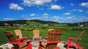 A view of a grassy landscape with houses in the distance, a fire pit, and three wooden Adirondack chairs.
