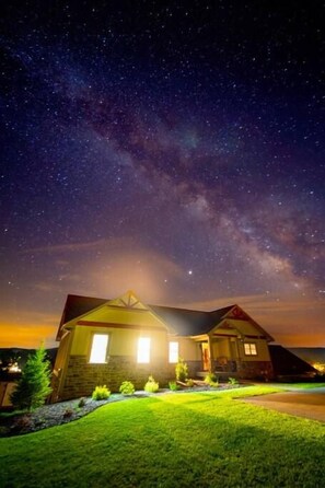 A house with a well-lit exterior, a clear view of the night sky, and a grassy lawn.