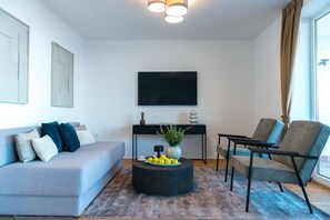 Uma sala de estar moderna com um sofá cinza, uma mesa de centro redonda preta com frutas, uma poltrona cinza e uma escrivaninha preta com uma TV de tela plana.