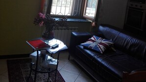 Una sala de estar con un sofá de cuero negro, una mesita con un jarrón de flores, un radiador y una ventana con cortinas.