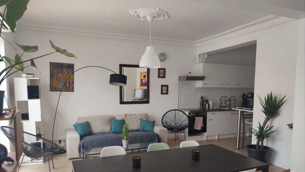 Una sala de estar moderna con una mesa de comedor negra, sillas blancas y verdes, un sofá con cojines color turquesa y una silla negra. Hay una zona de cocina con armarios blancos y una lámpara colgante.