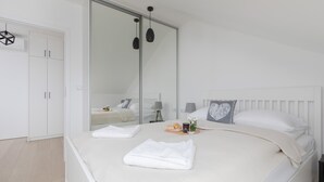 Un dormitorio con cama, cortinas blancas, armario, espejo y mesita de noche.