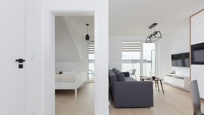 Una sala de estar moderna con un sofá, un comedor con mesa y sillas, un televisor montado en la pared, y una cama en la habitación contigua.