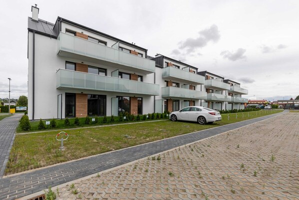A modern multi-story residential building with glass balconies, a paved driveway, and a car parked in front.