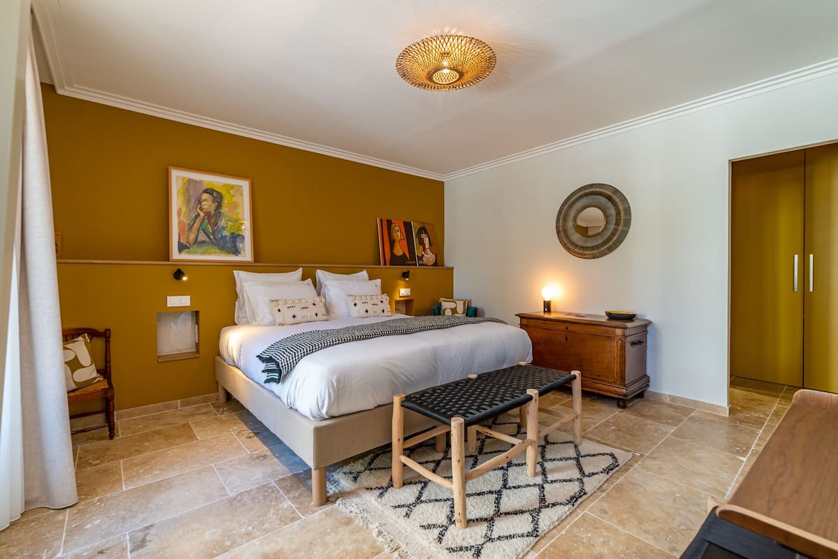 Quarto com cama, mesas de cabeceira, um banco, espelho fixado na parede e obras de arte.