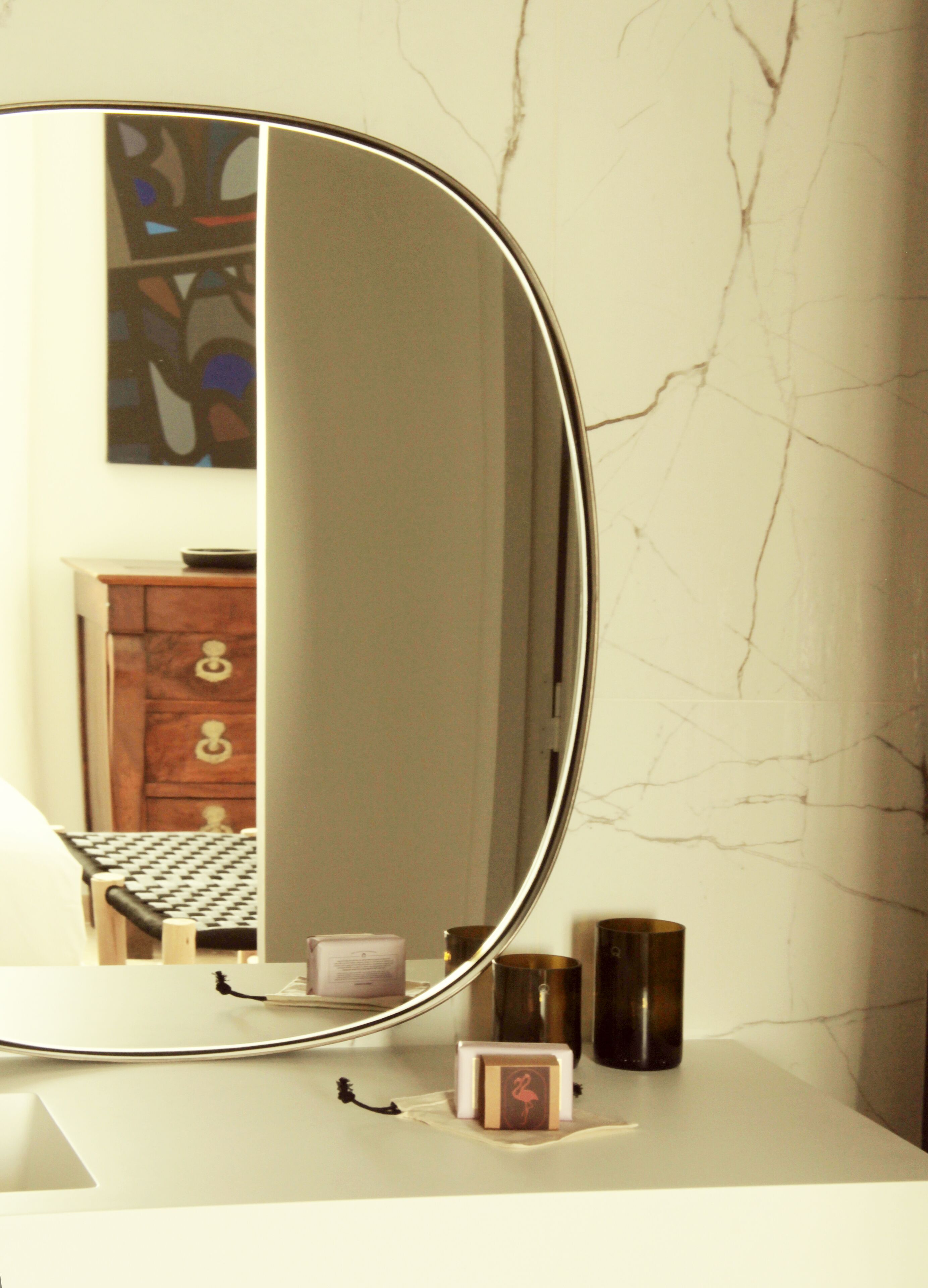 Un baño con pared de mármol, un espejo redondo, un mueble de madera y una pequeña estantería con artículos de aseo.