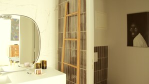 Une salle de bain avec un meuble-lavabo blanc, un miroir rond, une échelle en bois appuyée contre un mur carrelé et un tableau encadré accroché au mur.