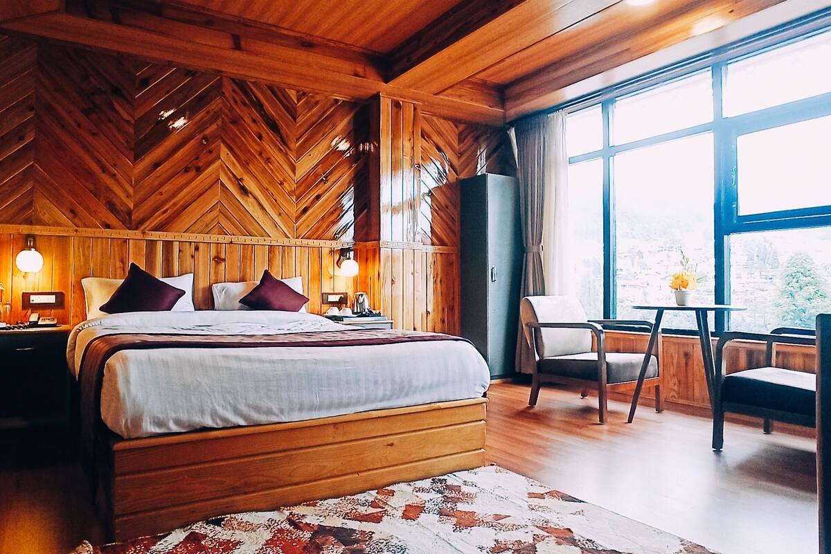 A wooden cabin bedroom with a bed, nightstands, a chair, a table, and a large window offering a view of the outdoors.