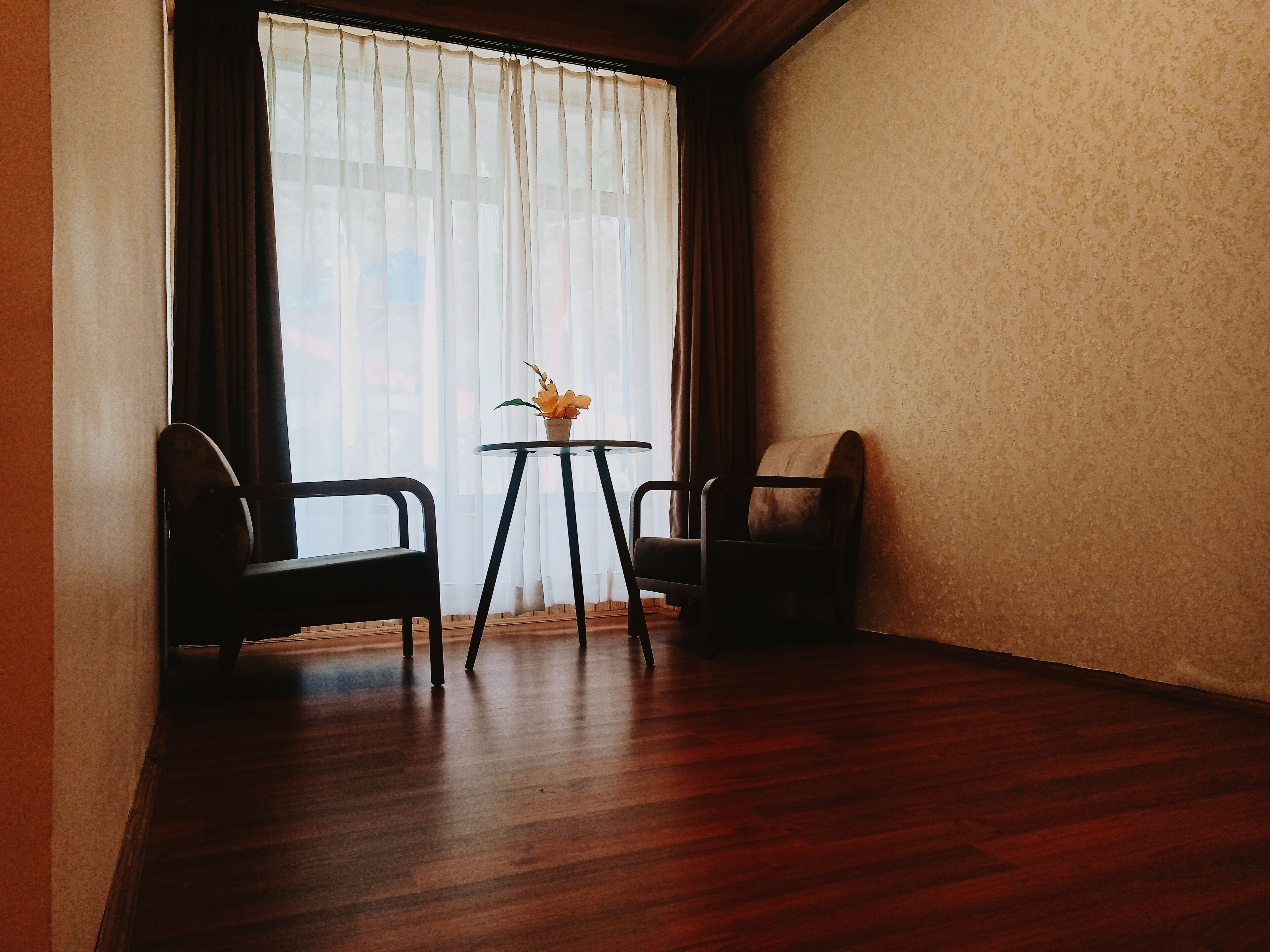 A room with two chairs, a small table with a flower, and a large window with sheer curtains.
