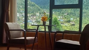 Habitación con un ventanal grande que ofrece vistas a un paisaje montañoso. Hay dos sillas y una mesita con una planta en maceta.