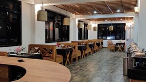Un restaurante con mesas y sillas de madera, ventanas amplias y lámparas colgantes.