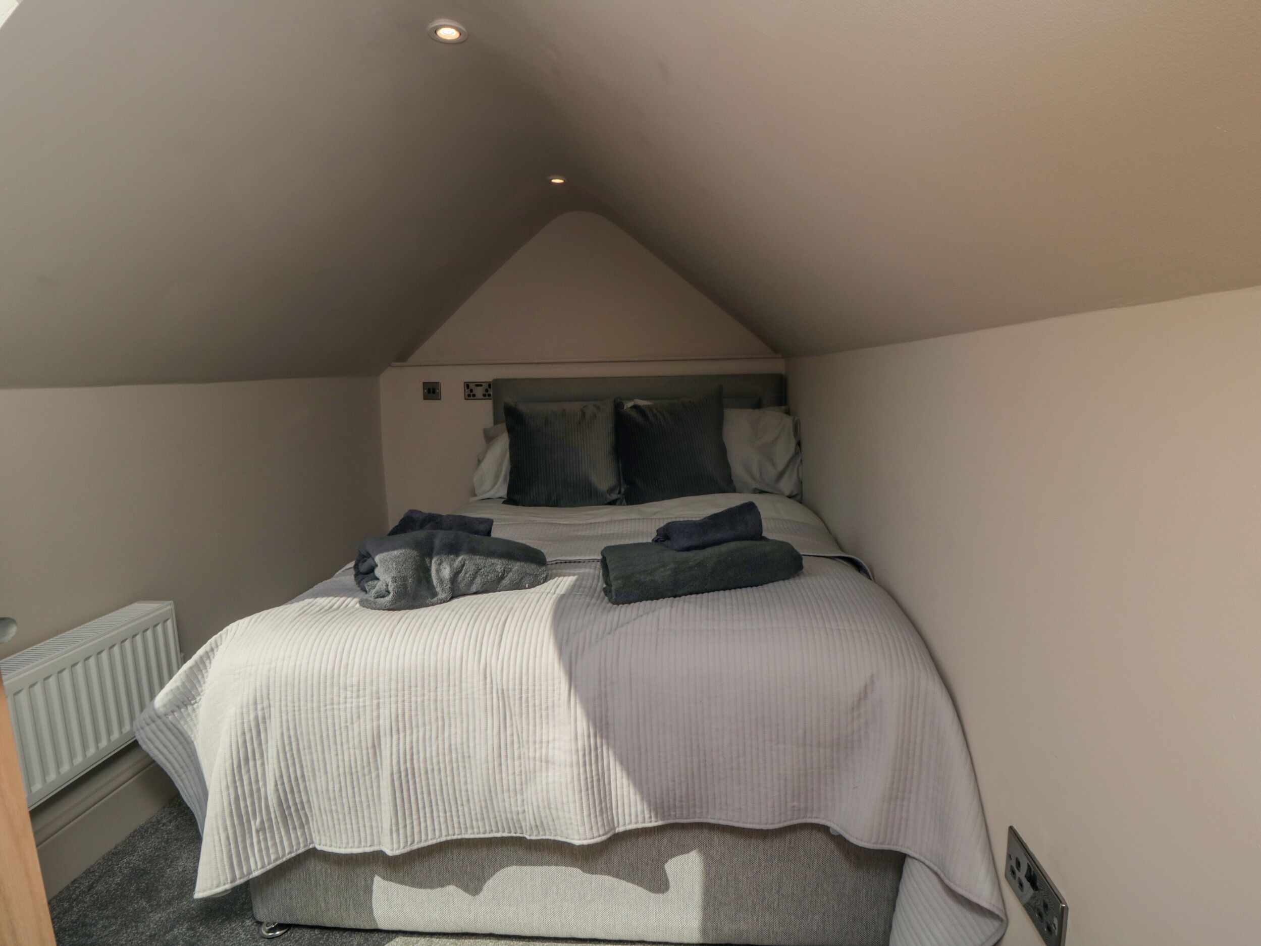 Une chambre avec un plafond en pente, un lit recouvert d’une couette à rayures grises et blanches, deux oreillers et une couverture pliée posée sur le lit.