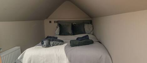 Une chambre avec un plafond en pente, un lit recouvert d’une couette à rayures grises et blanches, deux oreillers et une couverture pliée posée sur le lit.
