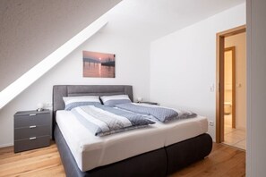 Ein modernes Schlafzimmer mit schrägen Wänden, einem Bett mit grauem Kopfteil, einem Nachttisch und einer Tür zu einem anderen Raum.