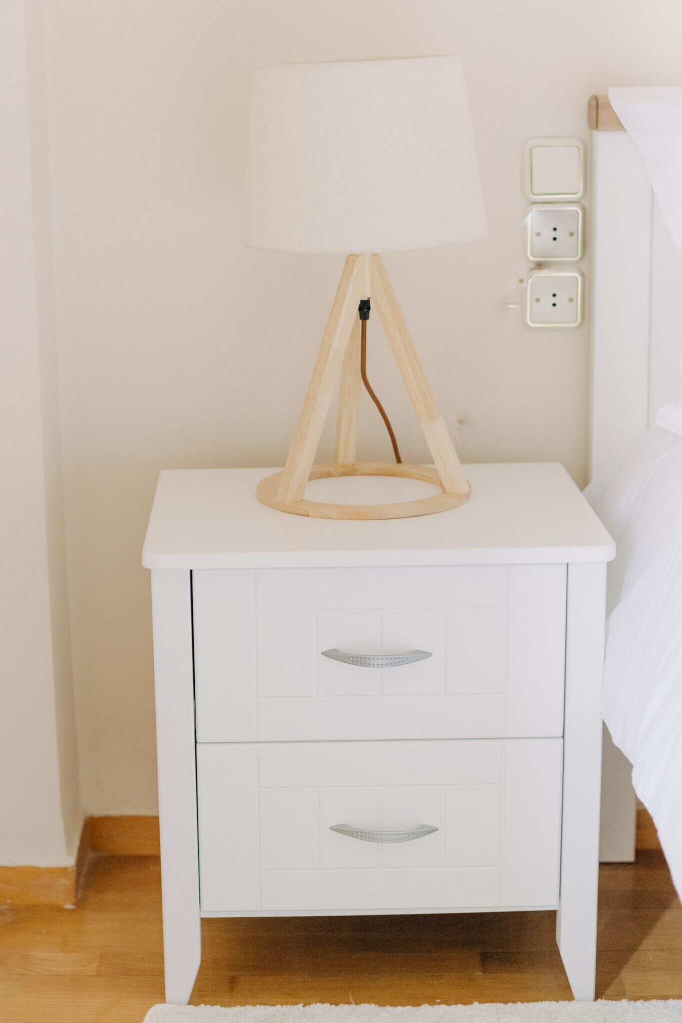 Een witte nachtkastje met een lamp, twee lades en een stopcontact aan de muur.