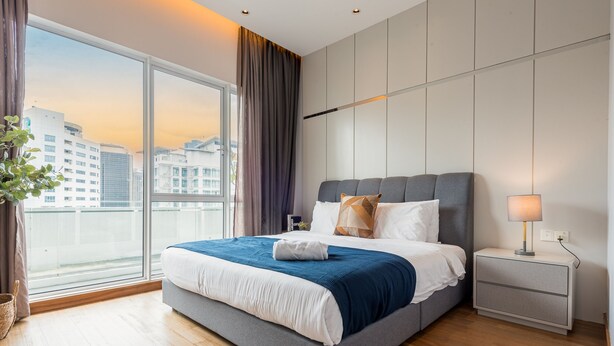 Ein modernes Hotelzimmer mit einem großen Bett, einem Nachttisch, einer Lampe und Blick auf die Stadt durch große Fenster.