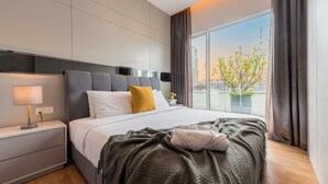 Ein modernes Hotelzimmer mit einem großen Bett, einem Nachttisch mit Lampe und Blick auf eine Stadtlandschaft durch ein großes Fenster.