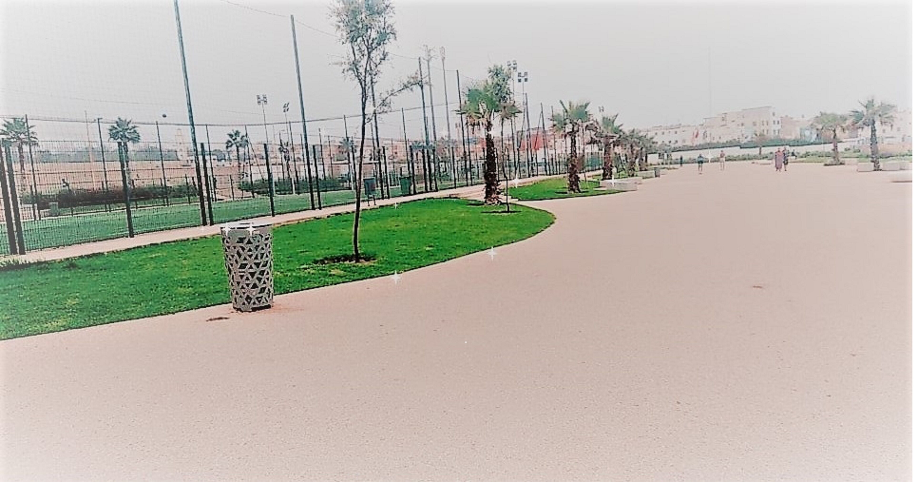 A paved pathway with a grassy area and trees, a patterned trash bin, and a fence in the background.
