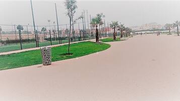 A paved pathway with a grassy area and trees, a patterned trash bin, and a fence in the background.