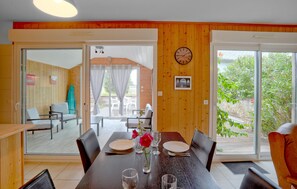 A dining area with a wooden table set for two, a clock on the wall, and a view of the outdoors through large sliding glass doors. - 2 bedroom beach front home in Créances (Créances)