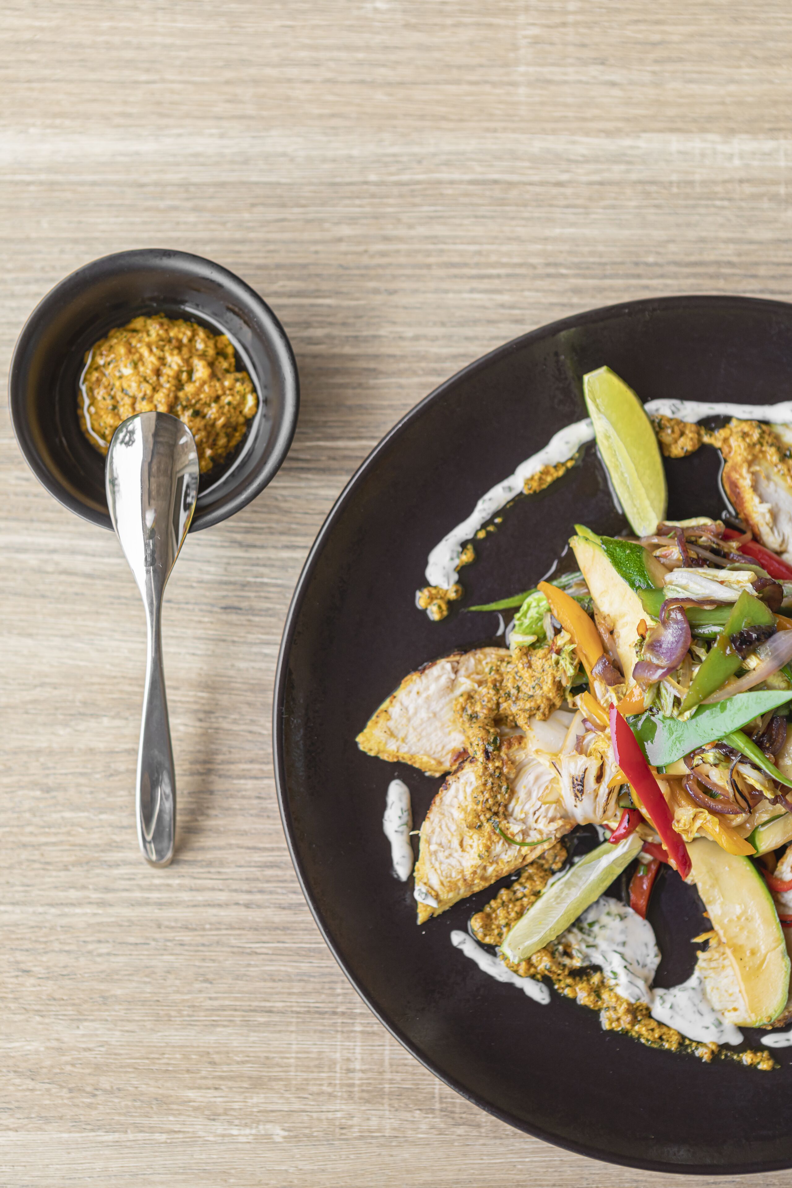 A black plate with grilled chicken, vegetables, and a lime wedge.