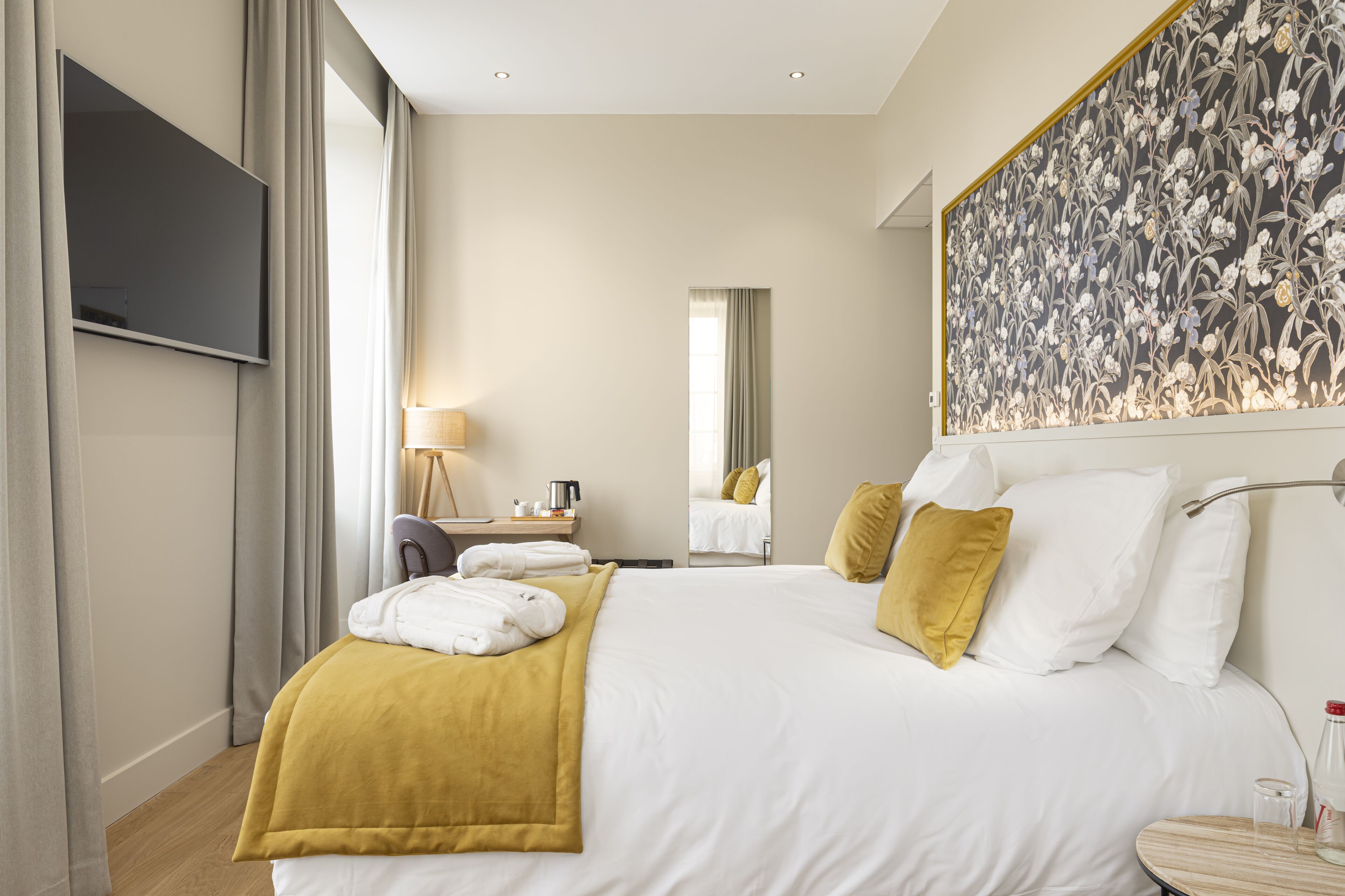 A hotel room with a large bed, a flat-screen TV, a desk with a lamp, and a large floral artwork on the wall.
