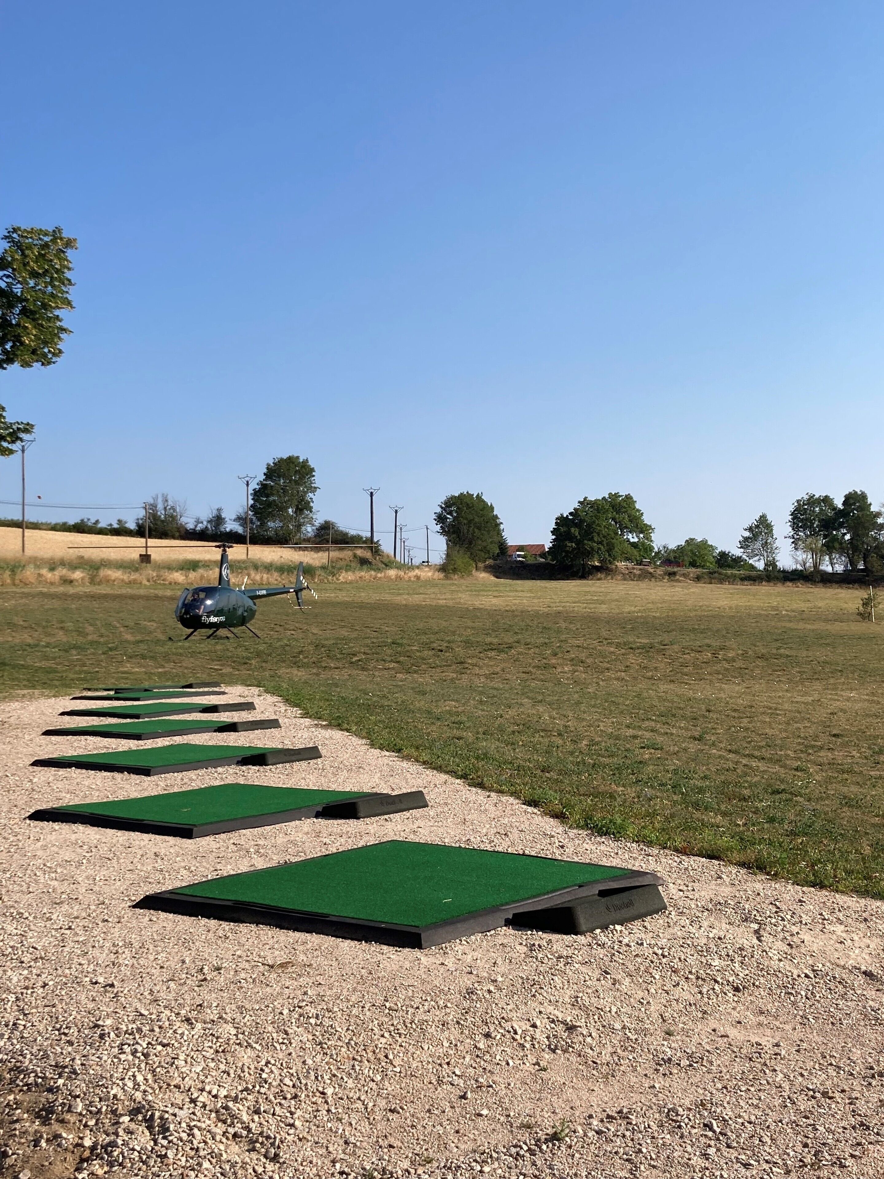 Campo de prácticas de golf con múltiples esteras verdes y un helicóptero al fondo.