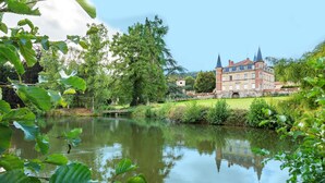 A large, historic building with multiple towers, surrounded by lush greenery and a calm lake.