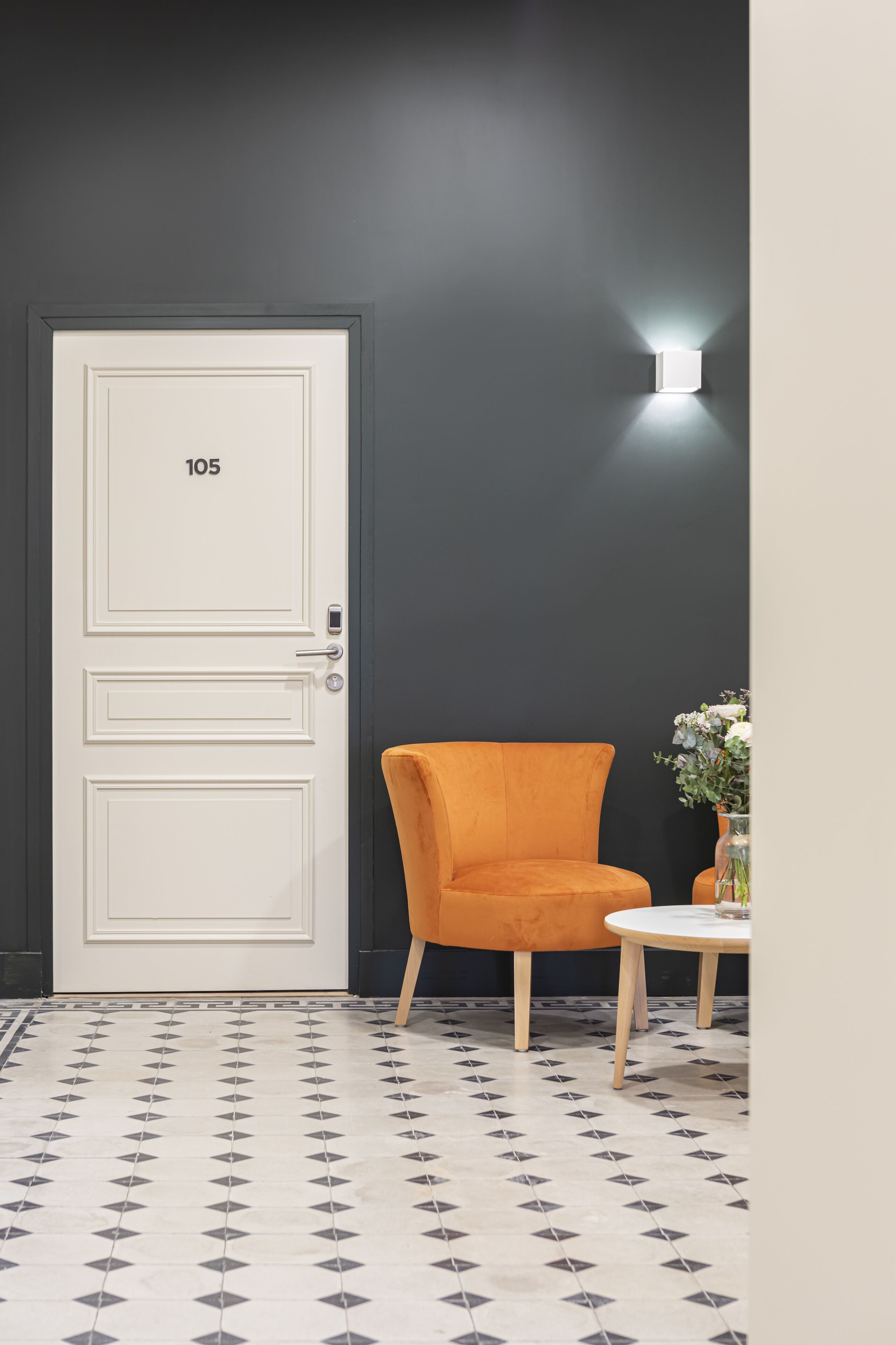 A room with a white door labeled "105," an orange chair, a small table with a vase of flowers, and a wall-mounted light fixture.