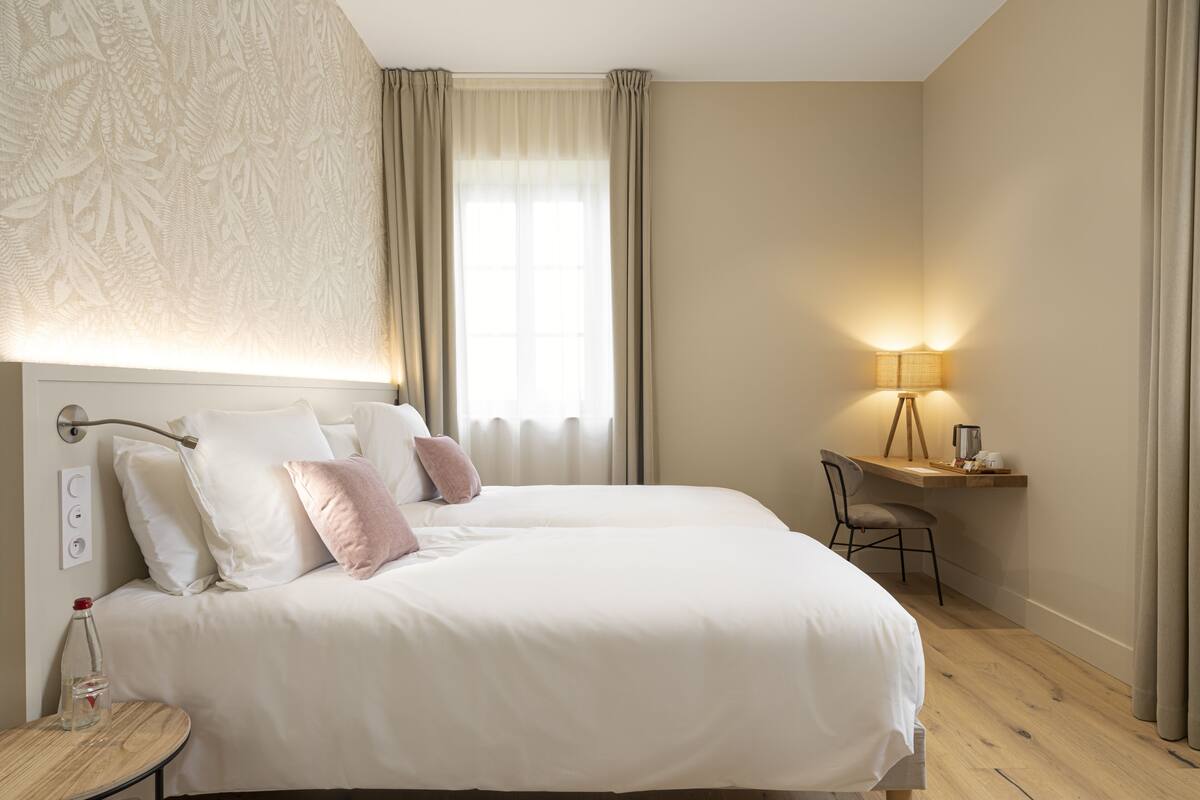 A hotel room with a large bed, a desk with a lamp, a chair, and a small table with a bottle.